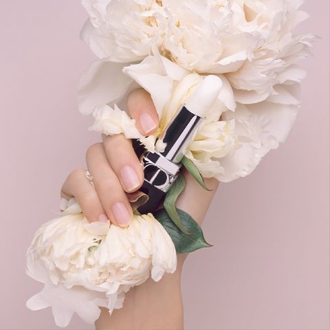 Una mano con uñas manicuradas sostiene un lápiz labial blanco, rodeada de grandes flores de peonía blanca, creando una atmósfera suave y elegante. El fondo es de color rosa claro, complementando la estética delicada.