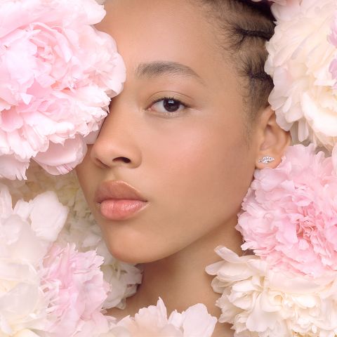 Una mujer está parcialmente rodeada de grandes peonías rosas y blancas. Su rostro está suavemente iluminado, mostrando un maquillaje natural y labios brillantes. En su oreja visible lleva un pequeño y elegante pendiente. El tono general de la imagen es suave y sereno.
