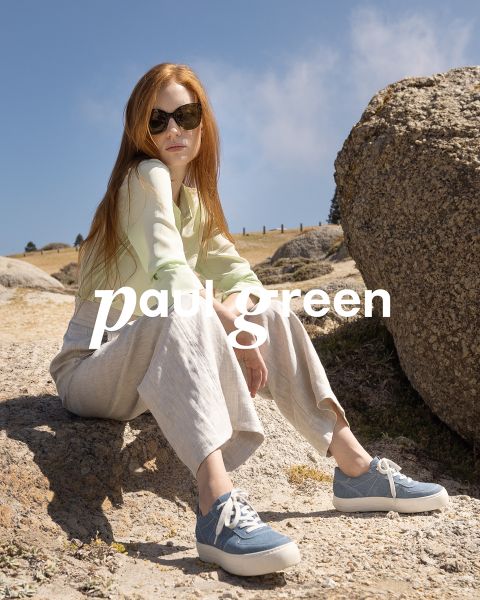 Una persona con pelo largo y rojo, gafas de sol, una blusa verde claro, pantalones beige y zapatillas azules está sentada en un terreno rocoso bajo un cielo azul. Las palabras "Paul Green" están superpuestas en la imagen.