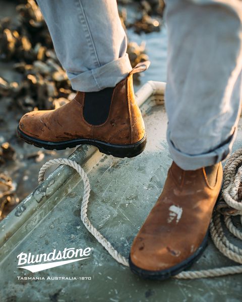 Una persona con botas Blundstone marrones desgastadas está de pie en un barco desgastado cerca de cuerdas, con agua y rocas borrosas al fondo.
