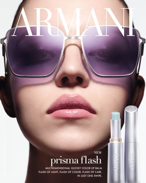 Primer plano de una mujer con gafas de sol extragrandes y labios rosados brillantes, con dos bálsamos labiales Armani Prisma Flash visibles en la parte inferior derecha.