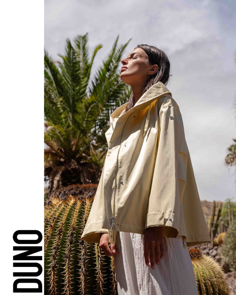 Una persona está de perfil, lleva una chaqueta amarilla clara y una falda blanca, tiene la cabeza inclinada hacia atrás y los ojos cerrados. Se encuentra en un entorno exterior con grandes cactus y palmeras. La palabra "DUNO" aparece verticalmente en el lado izquierdo de la imagen.