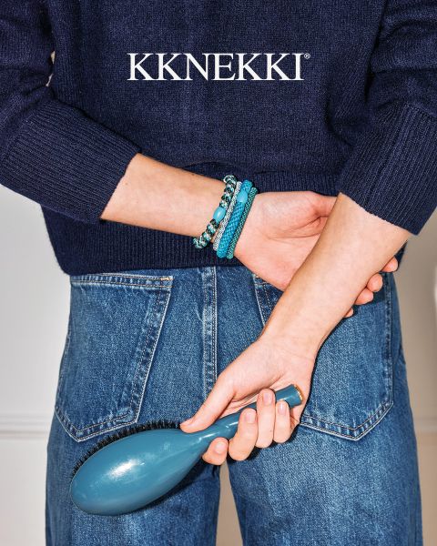 Una persona con un jersey azul marino y vaqueros sostiene un cepillo de pelo azul detrás de su espalda y lleva pulseras de cuentas turquesas. Arriba, el logotipo "KKNEKKI".