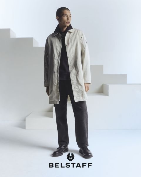 Una persona que lleva un abrigo claro sobre pantalones y zapatos oscuros está de pie frente a un fondo minimalista y geométrico. En la parte inferior de la imagen se ve el logo de "Belstaff".