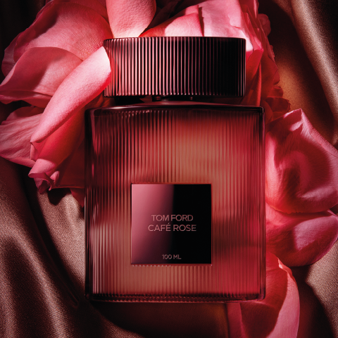 Una botella del perfume Tom Ford Café Rose está rodeada de pétalos de rosa sobre una tela sedosa de color bronce. La botella es rectangular con ranuras verticales y una etiqueta oscura cuadrada en el centro, que transmite lujo y elegancia.