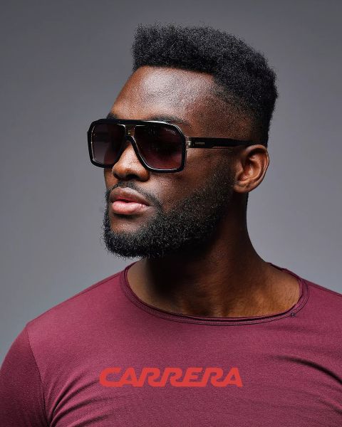 Hombre con grandes gafas de sol cuadradas y camisa burdeos con el logo de "CARRERA" mirando hacia un lado, sobre un fondo gris.
