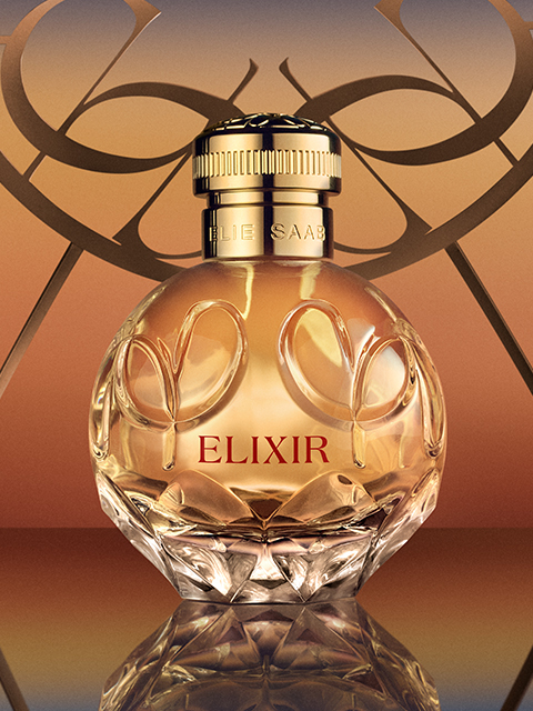 Un frasco de perfume de vidrio con la inscripción "Elie Saab Elixir" y un tapón dorado se encuentra frente a un cálido fondo degradado. El frasco redondo está adornado con grabados florales decorativos y refleja la luz, lo que le confiere un aspecto elegante y lujoso.