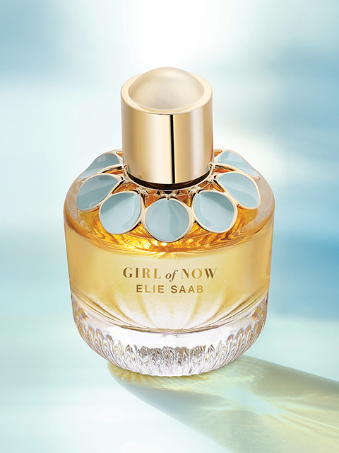 Una botella de vidrio de "Girl of Now" de Elie Saab se encuentra frente a un fondo suave y etéreo. La botella presenta detalles dorados y un motivo floral alrededor del cuello. La fragancia contiene un líquido cálido y ambarino, lo que contribuye a una apariencia lujosa y elegante.