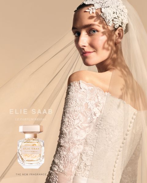 Novia con vestido de encaje y velo, con adorno floral en el cabello, mirando por encima del hombro. Su vestido proyecta una suave sombra con un patrón floral sobre un fondo beige. Se exhibe un frasco de perfume con el texto "Elie Saab Le Parfum Bridal".
