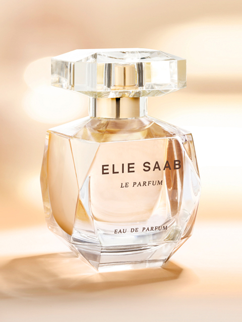 En un fondo suave y difuminado, se muestra un lujoso frasco de perfume con la inscripción "Elie Saab Le Parfum". El frasco es elegante, de vidrio transparente con un diseño geométrico facetado y contiene un líquido dorado.