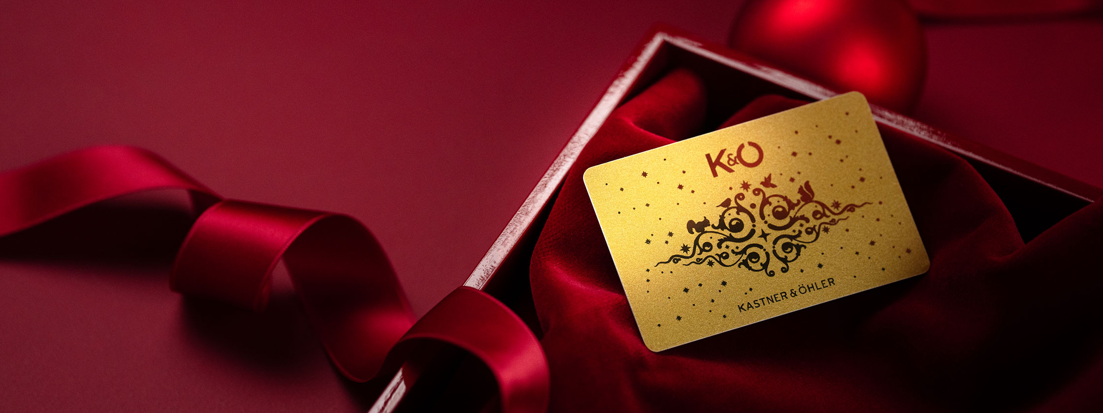 Eine goldene Geschenkkarte mit kunstvollen Mustern und dem Text „KO“ liegt auf einer roten Samtoberfläche in einer Schachtel. Daneben befinden sich ein rotes Band und eine rote Kugel, alles vor einem leuchtend roten Hintergrund.