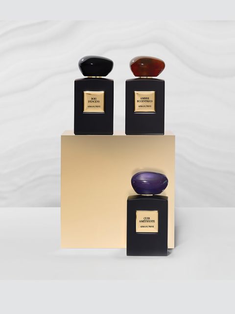Tres elegantes frascos de perfume sobre un pedestal beige. Cada frasco es rectangular y tiene un tapón ovalado único: uno en negro, uno en rojo y uno en púrpura. El fondo presenta un sutil patrón ondulado.