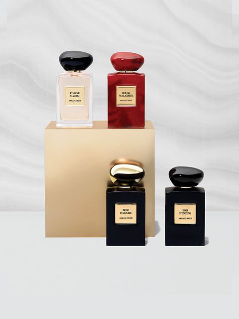 Cuatro elegantes frascos de perfume expuestos sobre una base dorada. Los frascos son negros, rojos y dorados y cada uno tiene un tapón a juego. Están dispuestos sobre un fondo suave y ondulado de color gris y blanco.