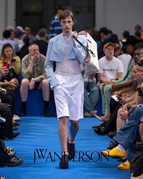 Una modelo en una pasarela azul lleva una camiseta corta azul claro, pantalones cortos blancos y zapatos de plataforma negros, y lleva un bolso blanco grande. Los espectadores sentados observan y toman fotos. El logo de J.W. Anderson se ve en el suelo.