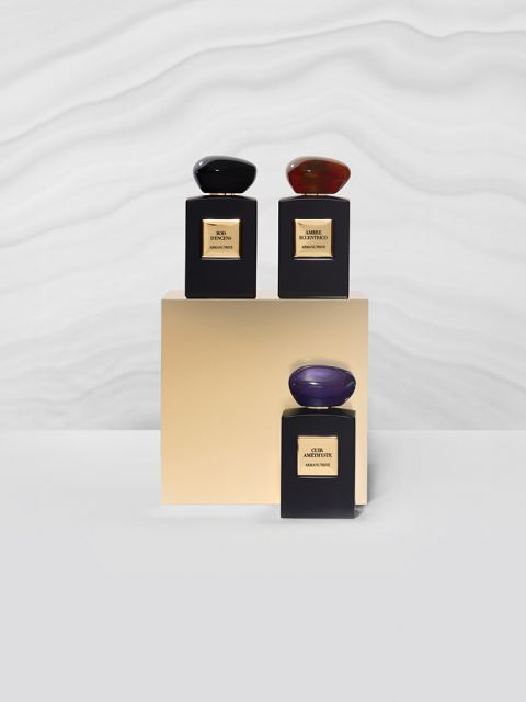Tres frascos de perfume sobre una base dorada frente a un fondo blanco ondulado y texturizado. Los frascos son negros con etiquetas doradas y tapas oscuras, lisas y ovaladas.