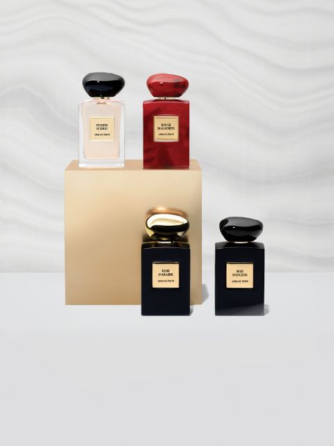 Cuatro frascos de perfume se exhiben sobre y alrededor de un pedestal beige. Los frascos son cuadrados con tapas redondeadas y en colores negro y rojo. Una pared blanca ondulada y texturizada sirve de fondo.