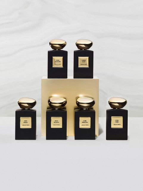 Se exhiben seis elegantes frascos de perfume con lisas tapas doradas. Están dispuestos sobre y alrededor de un bloque de oro y llevan varias etiquetas con las inscripciones "OUD", "SNAKE" y "OUR", cada una con intrincados diseños en negro y dorado sobre un suave fondo gris.