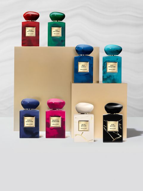 Ocho frascos de perfume en varios colores, incluyendo rojo, verde, azul, rosa y negro, están dispuestos sobre pedestales beige frente a un fondo claro. Cada frasco tiene una etiqueta con letras doradas.