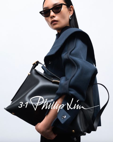 Persona con gafas de sol, abrigo oscuro y un bolso negro grande frente a un fondo sencillo. La superposición de texto dice "3.1 Phillip Lim" en escritura estilizada.