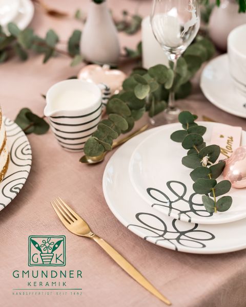 Una mesa bellamente dispuesta con platos de cerámica decorativos con patrones abstractos de líneas negras. Hojas de eucalipto descansan sobre los platos. Un tenedor dorado, una jarrita de crema a rayas y una taza estampada complementan el elegante mantel rosa.