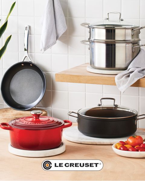 Una escena de cocina con una olla de cocción lenta roja, una sartén antiadherente, un juego de ollas apilables de acero inoxidable, una olla negra y un cuenco de tomates cherry sobre una encimera de madera clara frente a un protector contra salpicaduras de azulejos blancos. El logotipo de Le Creuset es visible.