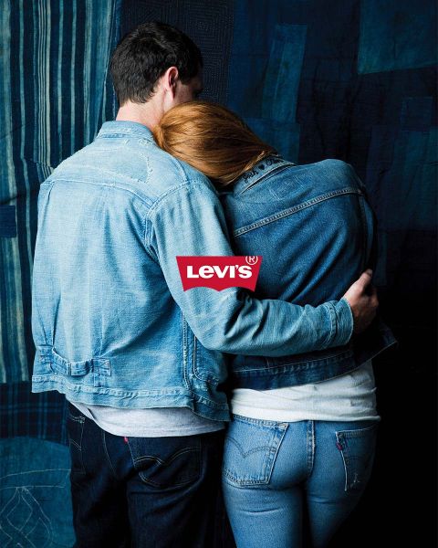 Hombre y mujer abrazándose frente a un fondo de mosaico azul, ambos con chaquetas y pantalones vaqueros. Encima de ellos, un logo rojo de Levi's destaca claramente.