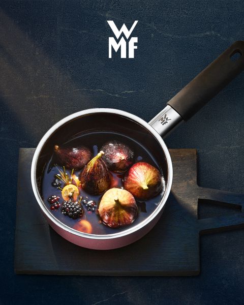 Una olla llena de higos, moras y guarniciones aromáticas está sobre una tabla de madera oscura. Encima de la olla se ve el logo de WMF. El fondo es una superficie oscura texturizada.