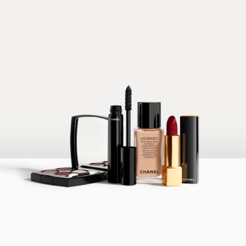 Una colección de productos de maquillaje de Chanel, que incluye un set compacto de sombras de ojos, una máscara de pestañas, una base líquida en un frasco de vidrio y un lápiz labial rojo oscuro en un estuche dorado y negro, dispuestos sobre una superficie blanca.