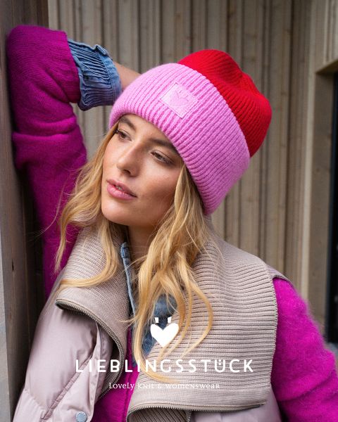Una mujer con un gorro de lana rosa rojizo, un suéter rosa claro, un chaleco acolchado beige y una camisa vaquera está de pie frente a una pared de madera. Parece pensativa y tiene la cabeza apoyada en la mano. El texto dice "PIEZA FAVORITA".