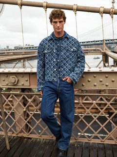 Un hombre con una chaqueta vaquera estampada y vaqueros azules está de pie en un puente con barandilla metálica. Al fondo se ven un río y otro puente en un día nublado.