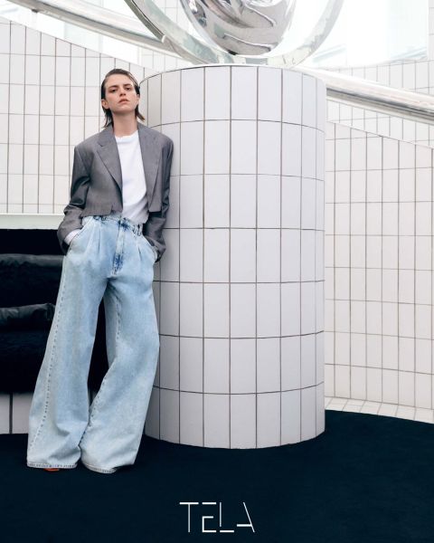 Persona apoyada en una columna con azulejos blancos, vistiendo un blazer gris, una camiseta blanca y vaqueros azul claro de gran tamaño. El suelo es negro y el entorno es minimalista con una decoración moderna. Abajo está la palabra "TELA".