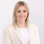 Una mujer con cabello largo y rubio sonríe a la cámara. Lleva un blazer claro sobre una blusa blanca. El fondo es de un blanco sencillo, transmitiendo una apariencia limpia y profesional.