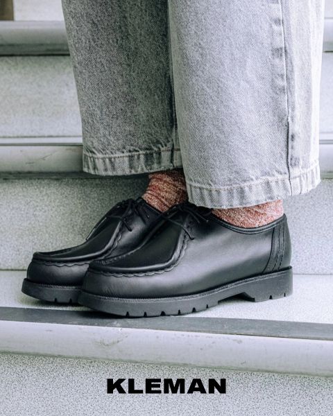 Primer plano de una persona con zapatos de cuero negros, calcetines con estampado rosa y vaqueros anchos de color claro, de pie en una escalera gris. El nombre de la marca "KLEMAN" aparece en la parte inferior de la imagen.