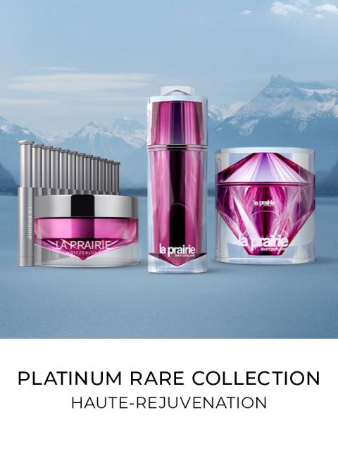 Una colección de productos para el cuidado de la piel Platinum Rare de La Prairie, que incluye una crema, un sérum y un aplicador. Tienen un elegante envase en color púrpura y plata con un fondo de montañas nevadas y un lago. Texto: "Platinum Rare Collection, Alta Rejuvenecimiento".