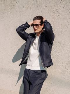 Un hombre con gafas de sol, una chaqueta azul marino y pantalones a juego está de pie con las manos en la cabeza frente a una pared clara y texturizada.