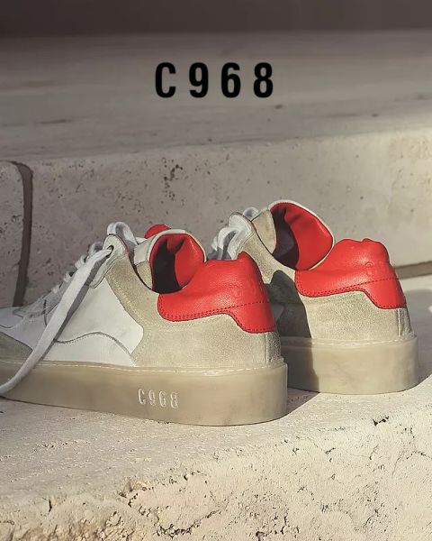 Un par de zapatillas deportivas elegantes sobre una superficie de piedra. Tienen partes superiores blancas y grises con fuertes detalles en rojo en el talón. El número "C968" aparece en el escalón del fondo y en las suelas de los zapatos.