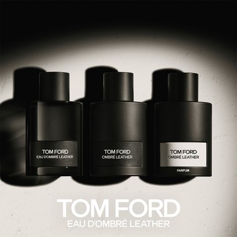 Tres frascos negros de perfume Tom Ford con las etiquetas "Eau d'Ombre Leather", "Ombre Leather" y "Parfum" están alineados. Proyectan sombras sobre una superficie clara, debajo está escrito en negrita "TOM FORD EAU D'OMBRE LEATHER".