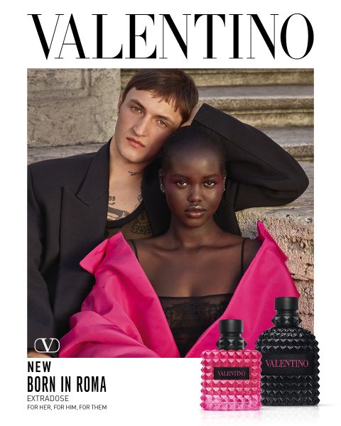 Un hombre y una mujer posan juntos en escalones de piedra. El hombre lleva un abrigo negro, la mujer un llamativo conjunto rosa con los hombros descubiertos. En la parte inferior derecha se ven dos frascos de perfume de Valentino. Encima pone "VALENTINO".