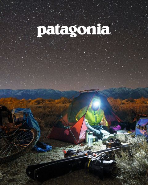 Una persona con linterna frontal sentada en una pequeña tienda iluminada bajo un cielo estrellado junto a una fogata. Cerca se ven bicicletas, equipo para exteriores y arriba un logo de Patagonia. Al fondo se ven un paisaje árido y una cadena montañosa.