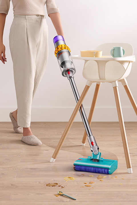 Una persona usa una aspiradora inalámbrica para limpiar cereal derramado en un piso de madera junto a una silla alta blanca. Una taza y un tazón están en la bandeja de la silla alta. La persona lleva pantalones claros y zapatillas.