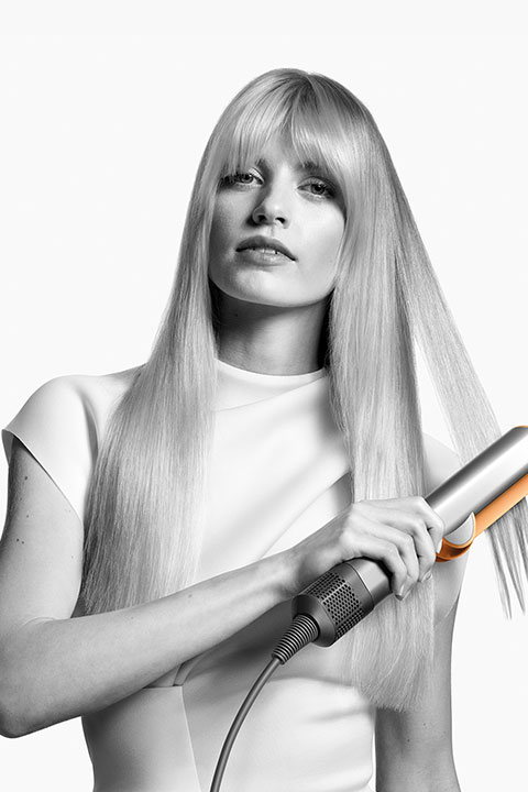 Mujer con cabello largo y rubio usando una plancha, vistiendo una blusa blanca y mirando hacia adelante. Imagen en blanco y negro de alto contraste.