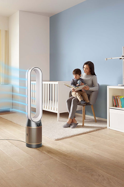Una mujer está sentada en una silla leyendo a un niño en una habitación infantil con una cuna, una estantería y una lámpara. En primer plano, un purificador de aire con un diseño elegante y moderno emite corrientes de aire. La habitación tiene suelos de madera clara y paredes azules suaves.
