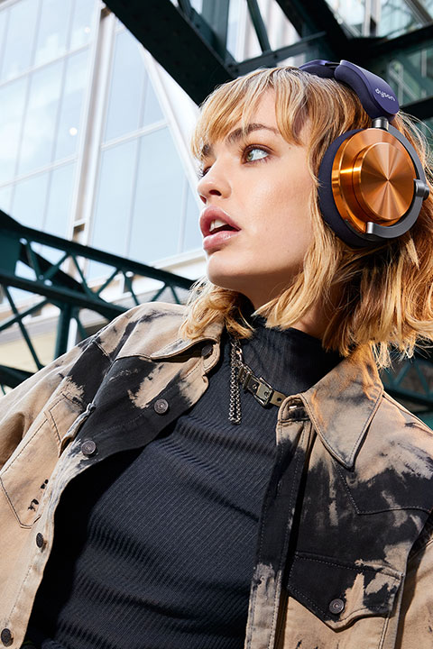 Una persona con cabello rubio corto lleva unos grandes auriculares color cobre y una chaqueta batik sobre un jersey de cuello alto negro. Mira hacia un lado, en el fondo se ve una construcción industrial de vidrio y metal.