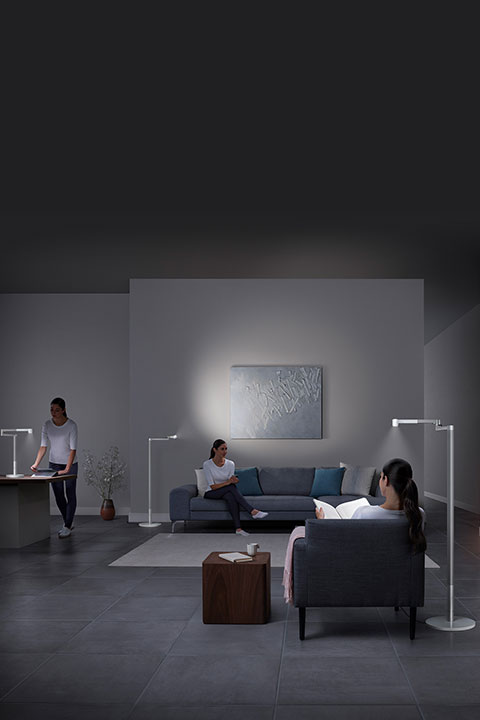 En una sala de estar con poca luz, tres personas están ocupadas en diferentes actividades: una está de pie en un escritorio, otra está sentada con un libro en un sofá y una tercera está sentada leyendo en una silla. La decoración moderna y las lámparas de pie iluminan la habitación.