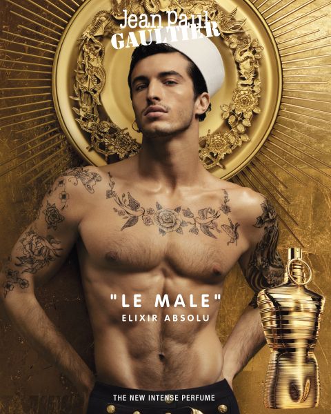 Un modelo masculino sin camisa con tatuajes lleva un gorro de marinero frente a un fondo dorado y anuncia el perfume "Le Male" Elixir Absolu de Jean Paul Gaultier. En la esquina inferior derecha hay un frasco de perfume dorado. El texto dice: "El nuevo perfume intenso".