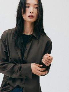 Mujer con pelo largo y negro, que lleva una camisa marrón holgada y una camiseta negra, mirando hacia un lado frente a un fondo blanco liso.