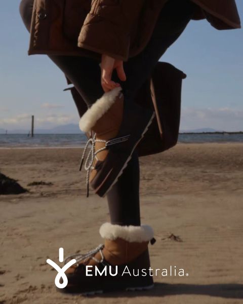 Una persona con botas EMU Australia marrones con forro de piel y un abrigo marrón está de pie en una playa de arena, levantando un pie detrás de ella. El logotipo de EMU Australia aparece en la parte inferior de la imagen.