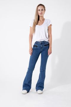 Una persona con una camiseta blanca y pantalones acampanados azules está de pie frente a un fondo blanco liso. Tiene el pelo largo y lleva zapatillas blancas. Está posando con una mano en el bolsillo y la otra en la cadera.