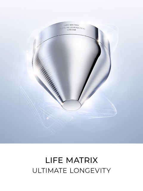 Recipiente cosmético futurista plateado con sutiles líneas brillantes alrededor, con la inscripción "Life Matrix Crema de Rejuvenecimiento Haute". El texto debajo dice "Life Matrix Longevidad Máxima" sobre un fondo blanco.