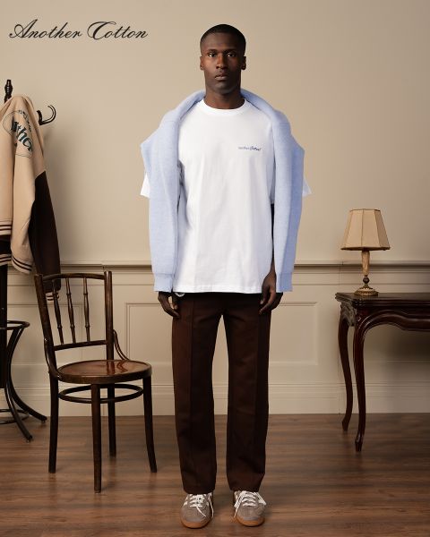 Una persona está de pie en una habitación, lleva un suéter azul claro sobre los hombros, una camiseta blanca y pantalones marrones. En la habitación hay una silla de madera, una lámpara de pie vintage y una chaqueta en un perchero. En la imagen se ve el texto "Another Cotton".